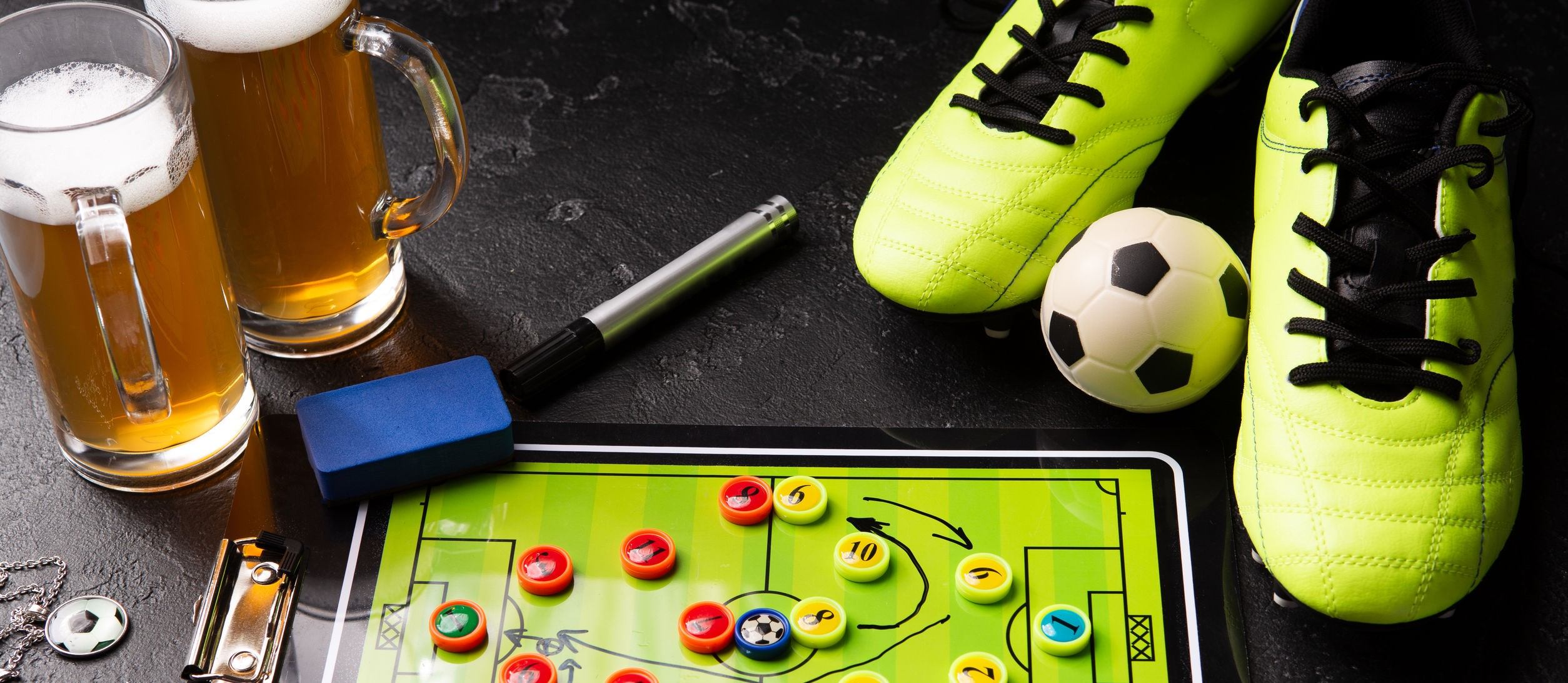Photo of two mugs of foam beer, table football, ball, football boots, pipe, rattle toy on black table