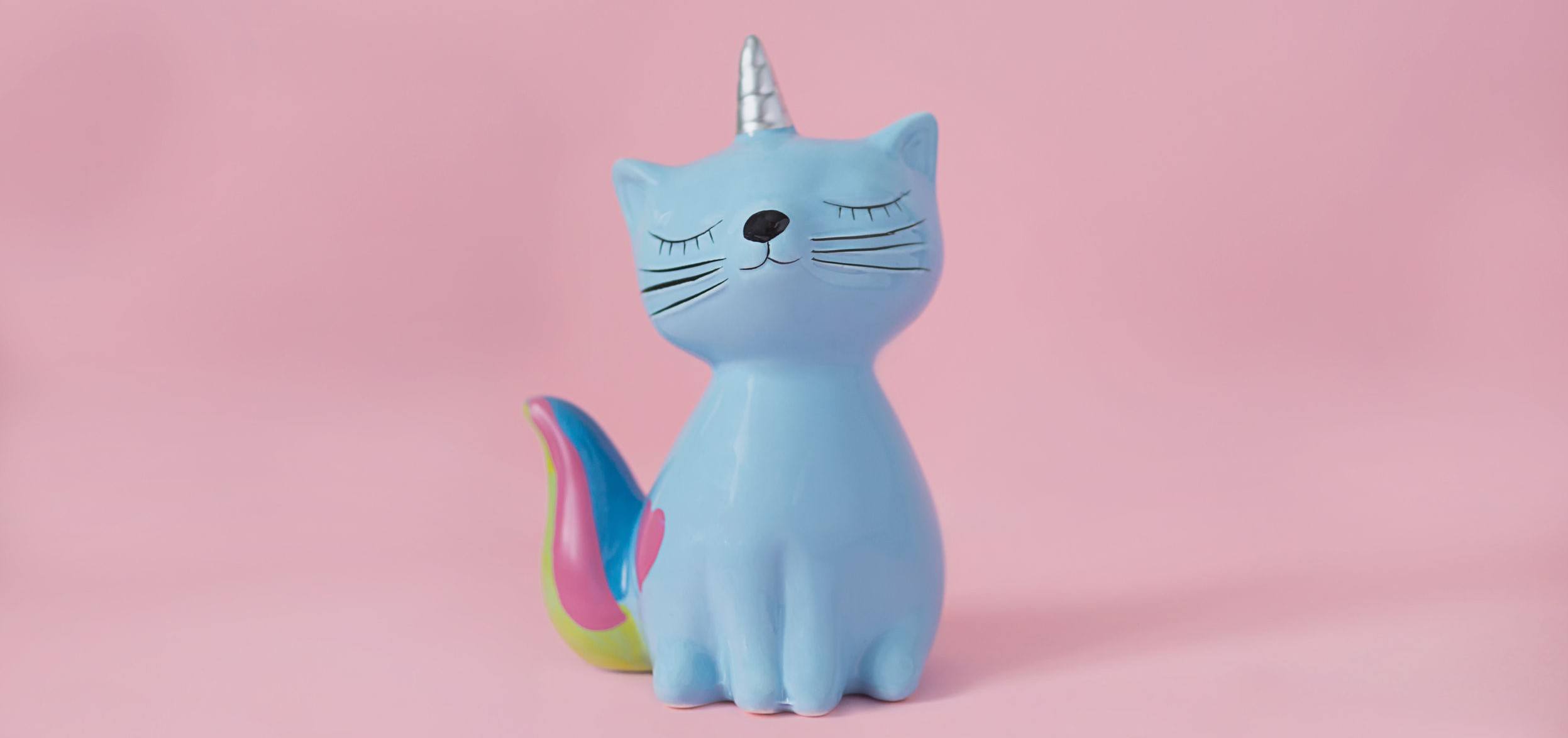 Picture of a ceramic souvenir toy moneybox kitten Korn blue with colorful rainbow tail with closed eyes and unicorn horn on pink background in natural light